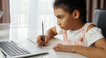 Fotografía de niña sentada, con papel y pluma en mano, frente a una computadora