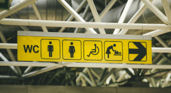 Imagen de señalización en un espacio público. De izquierda a derecha: WC, icono de hombre, icono de mujer, icono de persona usuaria de silla de ruedas, icono de cambiador de pañales, icono de una flecha señalando a la derecha. Foto por Paul Green para Unsplash