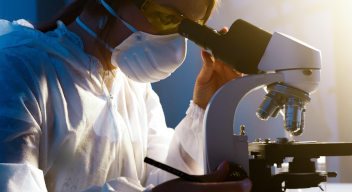Foto de una mujer de perfil. Trae puesta una bata médica y está viendo a través de un microscopio en un laboratorio. Foto por Artem Poder para Pexels.