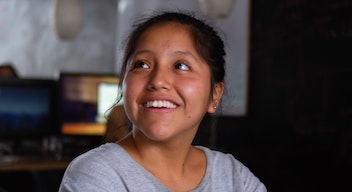 Una niña morena, de pelo negro y ojos cafés, de alrededor 10 años, sonríe mirando hacia la izquierda.
