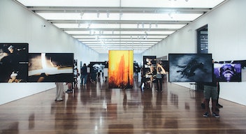 Fotografía de una sala de exposición en un museo. El piso es de madera. Del techo cuelgan cuadros de fotografías.