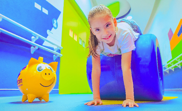 Niña sonriendo encima de un cilindro y al lado alcancía de cochinito